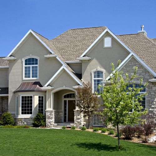 Front view of a large residential home with a green lawn, featuring an exterior finished with beige stucco and light-colored stacked stone veneer, arched entry, and architectural windows.