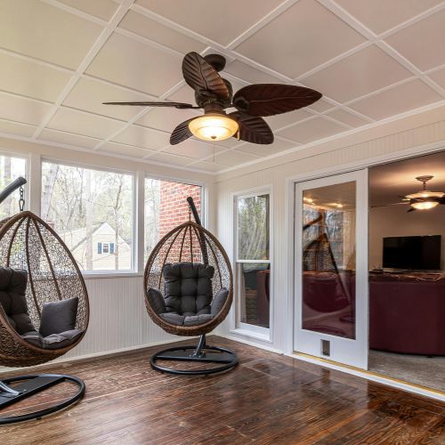 Finished sunroom or enclosed porch with dark stained wood flooring, white beadboard walls, two rattan hanging chairs, a detailed coffered ceiling, and a bronze leaf-style ceiling fan installation.