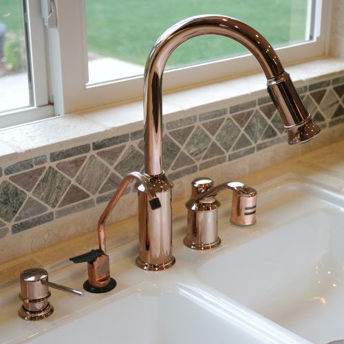 Close-up of a newly installed rose gold finish gooseneck pull-down kitchen faucet and soap dispenser, set on a white dual-basin sink with a decorative stone mosaic tile backsplash.