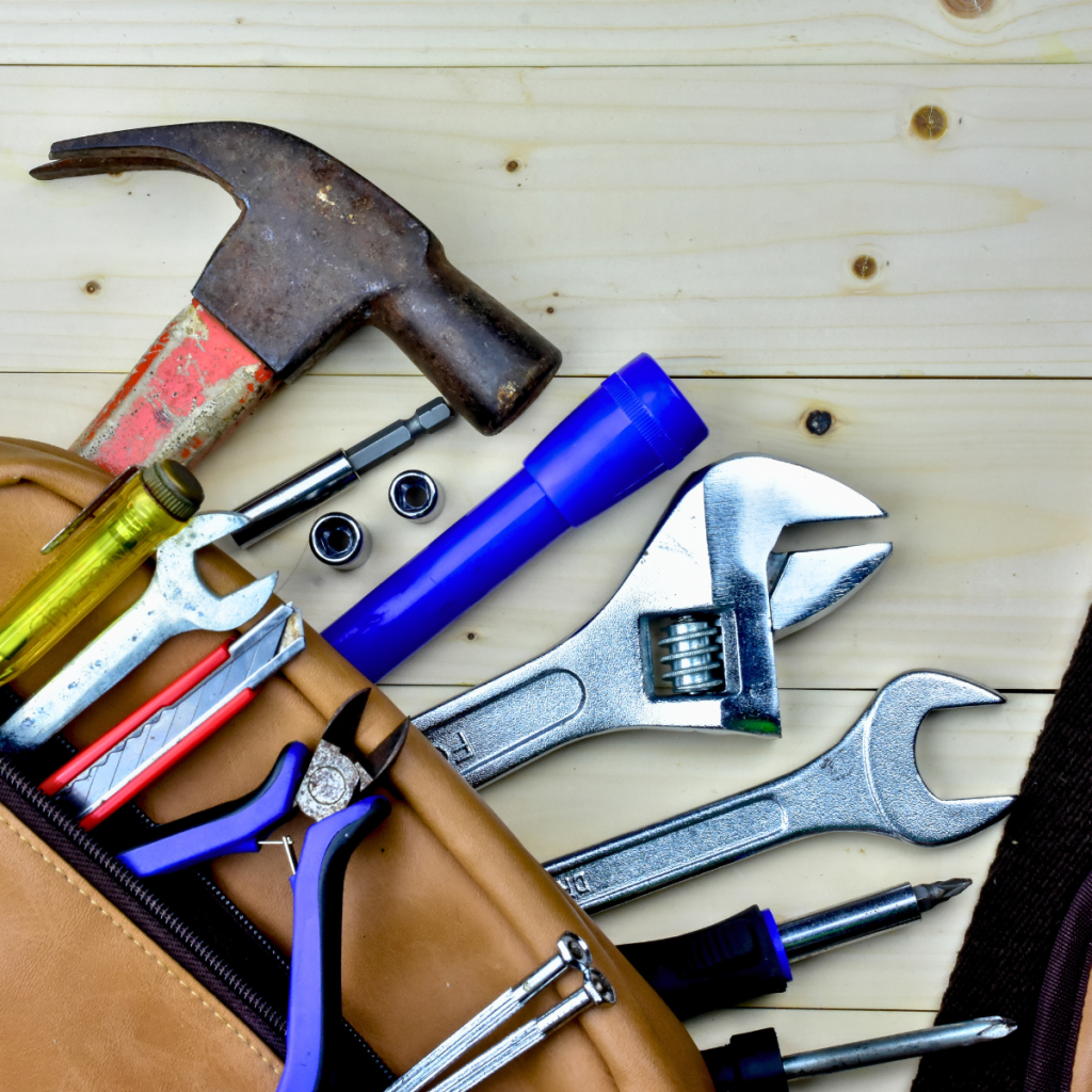 Overhead view of essential handyman tools, including a rust-tinged hammer, adjustable wrench, box wrench, screwdriver, flashlight, utility knife, and nails spilling out of a brown leather tool pouch onto a wooden surface.