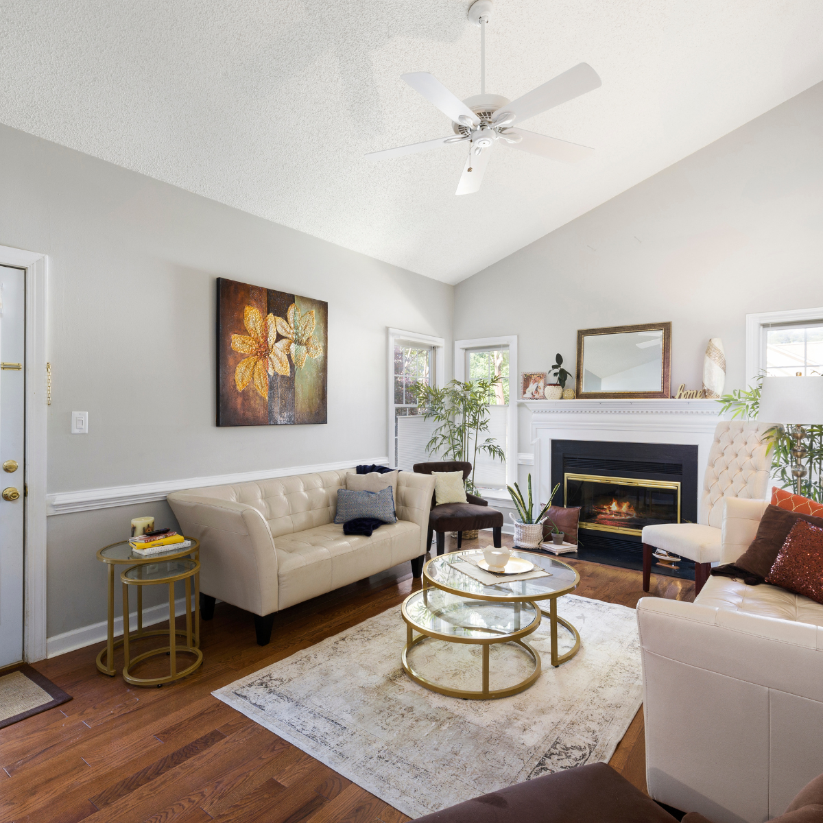 Cozy, fully decorated living room with vaulted ceiling, hardwood floors, white electric fireplace mantel, and white ceiling fan installation. Room features neutral furniture and wainscoting.