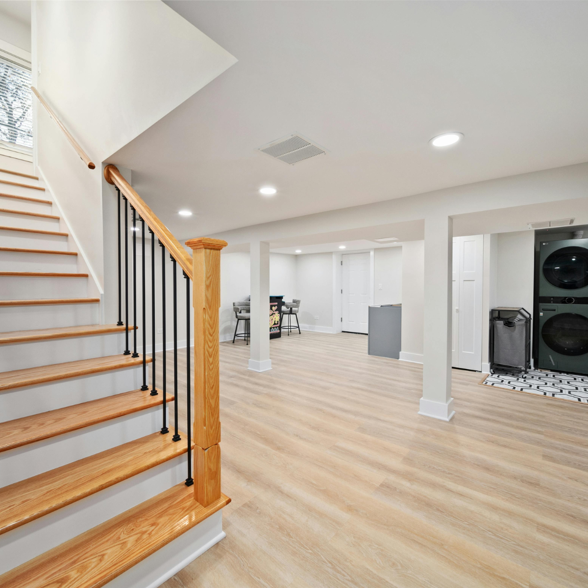 Bright, newly finished basement space featuring light wood-look laminate flooring, recessed lighting, white painted walls, a laundry area, and a staircase with natural wood treads, newel posts, and black metal balusters.