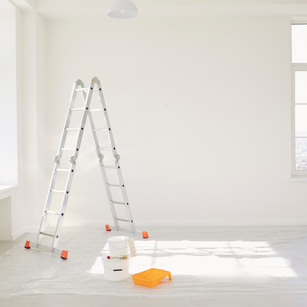 Minimalist, empty room with white walls and windows, covered in plastic sheeting on the floor for painting prep, featuring an aluminum A-frame ladder, paint cans, and a roller tray.