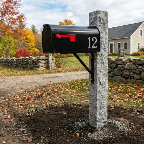 A sturdy grey granite mailbox post with a black metal mailbox and red flag, installed near a classic stone wall in a New Hampshire front yard.