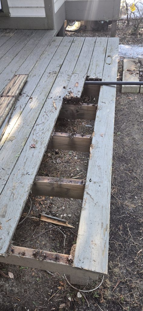 A pry bar resting on partially removed old wooden deck boards, showing the bare structural frame underneath before the new cedar installation.