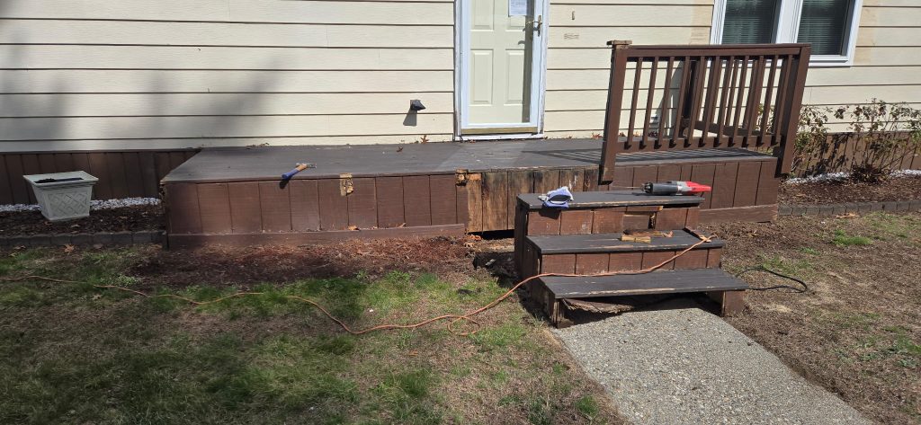Straight-on view of an unsafe, rotting front entry deck with detached wooden stairs and power tools during a demolition and replacement project in Goffstown, NH.