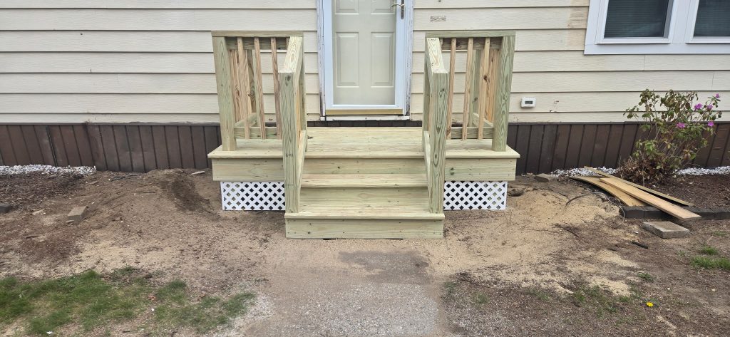 A newly built pressure-treated wood front entry landing and steps featuring sturdy railings and white lattice skirting, completed as a porch step replacement in Goffstown, New Hampshire.