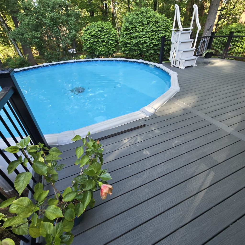 A beautiful new gray composite deck built around an above-ground pool, featuring black metal safety railings and white pool stairs, constructed by a custom deck builder in Londonderry, New Hampshire.