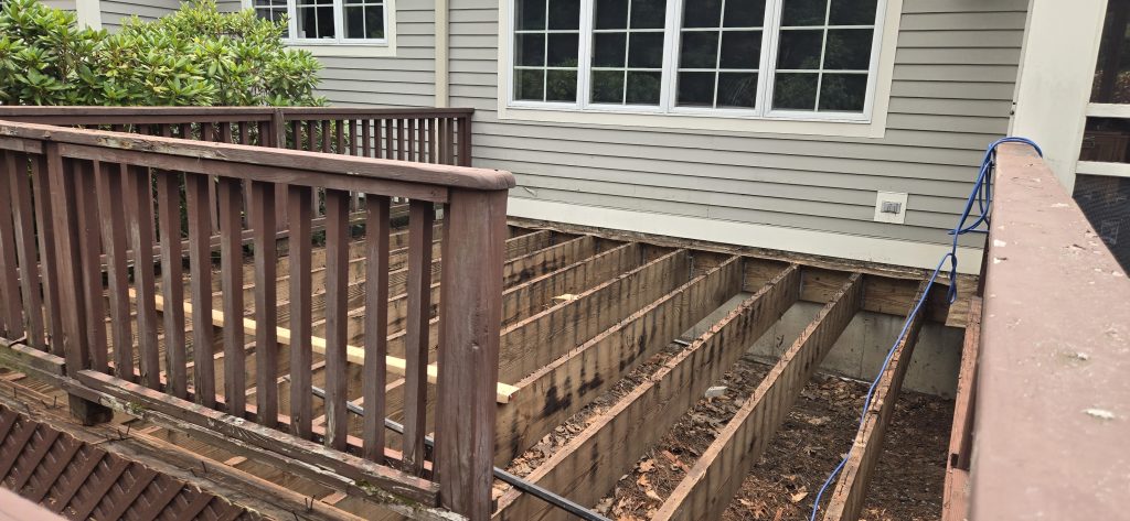 A residential deck repair and rot restoration project in Bow, New Hampshire, showing the old floorboards removed to expose the wooden joists and framing for structural inspection before resurfacing.