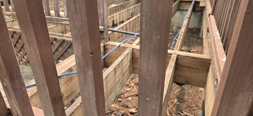 A close-up view through brown wooden railings showing exposed deck joists and framing during a deck rot repair and restoration project